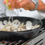 A chef squeezes a lemon over white clams cooking in a pan with white wine